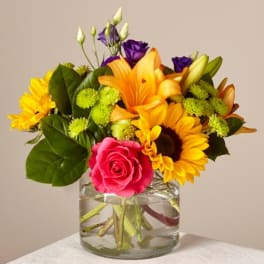 Mixed bouquet with sunflowers, roses, lilies, and purple blooms in a glass vase