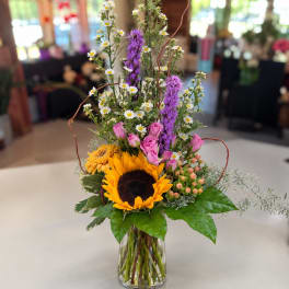 Mixed bouquet with sunflowers, pink roses, daisies, and purple spikes in a glass vase