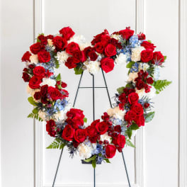 Heart-shaped floral wreath of red roses, white flowers, and blue accents on a stand