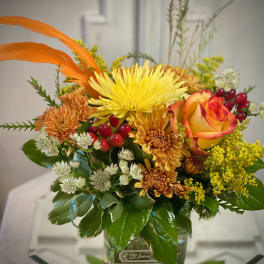 Orange and yellow floral arrangement in a glass vase with a rose and chrysanthemums