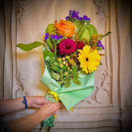 Handheld bouquet of orange, pink, and yellow flowers wrapped in mint paper