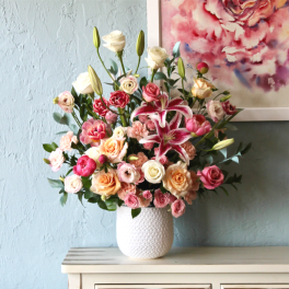 Large mixed bouquet of pink lilies and pastel roses in a white textured vase on a cabinet.