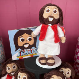Plush Jesus doll with a red scarf beside a matching book and smaller dolls