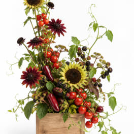 Mixed floral arrangement with sunflowers, dark daisies, berries, and tomatoes in a wooden box