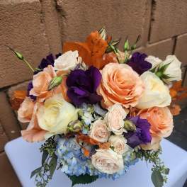 Compact arrangement of peach and cream roses, purple lisianthus, and blue hydrangea with orange leaves
