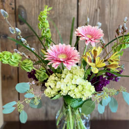 Tall mixed bouquet with pink gerbera daisies, green hydrangeas, and yellow blooms in a clear glass vase.