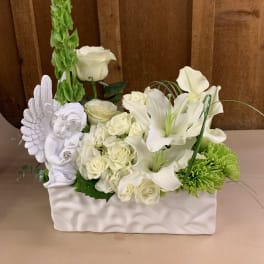 White floral arrangement with roses, lilies, and a cherub figurine in a white container