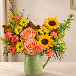 Bouquet of peach roses, sunflowers, and yellow daisies in a green pitcher vase