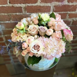 Round arrangement of blush roses and hydrangeas in a white vase against a brick wall.