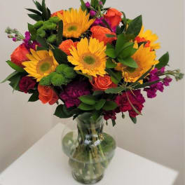 Bouquet of sunflowers, orange roses, and purple flowers in a clear glass vase