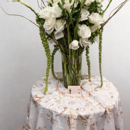 Tall white flower arrangement with roses and trailing green accents in a clear glass vase on a round table.
