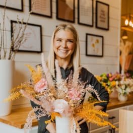 Florist holding a tall white vase with pink flowers and feathery golden dried accents in a shop.