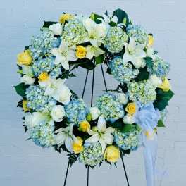 Large floral wreath with white lilies, yellow roses, and blue hydrangeas on a stand