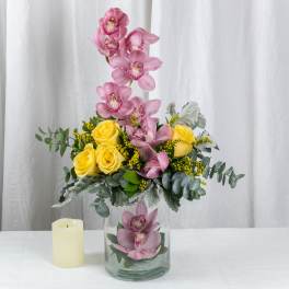 Tall arrangement of pink orchids and yellow roses in a clear glass vase beside a cream pillar candle