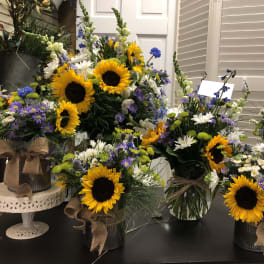 Several mixed flower arrangements with sunflowers and daisies in vases and tins