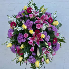 Large standing spray with pink lilies, roses, yellow roses, purple mums, and white accents on an easel.