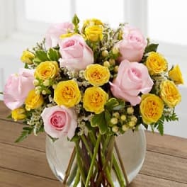 Round glass vase of pink roses and yellow spray roses with white filler flowers.