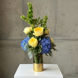 Yellow roses and blue hydrangeas in a gold vase with curly branches