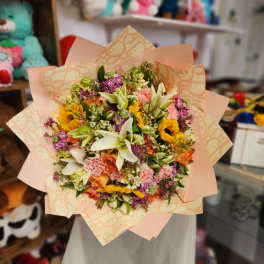 Mixed bouquet of lilies, sunflowers, carnations, and purple blooms in peach wrap