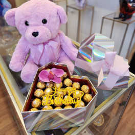Lavender teddy bear beside a heart-shaped chocolate box with a birthday topper