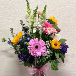 Mixed bouquet of pink, yellow, white, and purple flowers in a glass vase with a pink ribbon