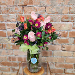 Pink roses and purple flowers arranged in a glass vase with a green ribbon.