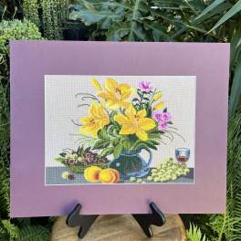 Framed floral needlepoint with yellow lilies and fruit on a stand