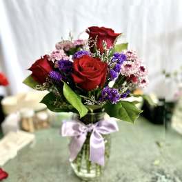 Red roses and purple filler flowers in a glass vase with a lavender ribbon