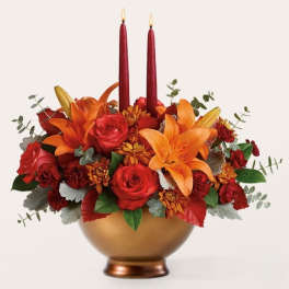 Red and orange floral arrangement in a gold bowl with two red taper candles