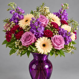 Bouquet of pink roses, purple flowers, and cream gerbera daisies in a purple vase