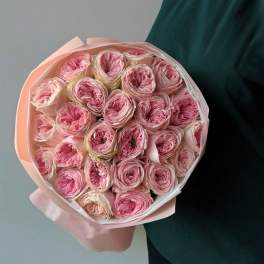 Round bouquet of tightly packed pink roses wrapped in pale peach paper, held by a person in dark clothing