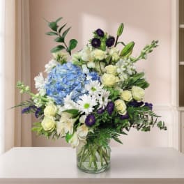 Mixed bouquet of blue hydrangeas, white daisies, and pale roses in a glass vase