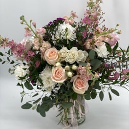 Tall arrangement of peach roses, white mums, and pink blooms in a glass vase with trailing ribbons