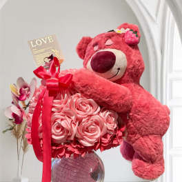 Pink teddy bear hugging a bouquet of pink roses in a glass vase