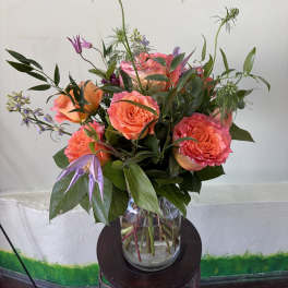 Vase arrangement of coral roses with purple accent flowers in a clear glass vase