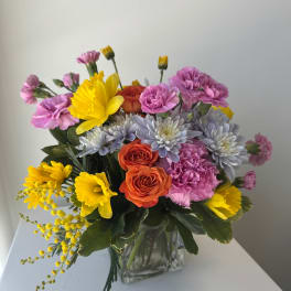 Mixed bouquet of yellow, pink, orange, and lavender flowers in a clear glass vase