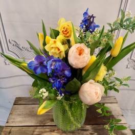 Mixed bouquet in a green glass vase with yellow, blue, and peach flowers