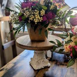 Basket-style arrangement of purple roses and white blooms with green accents in a brown container.