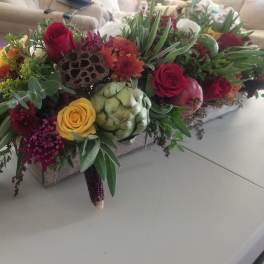 Low centerpiece of red and yellow roses, mums, protea, herbs, and artichoke in a rustic rectangular box