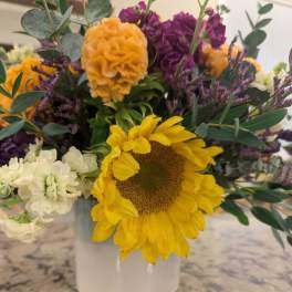 Bright arrangement with a large yellow sunflower, orange and purple blooms in a white vase
