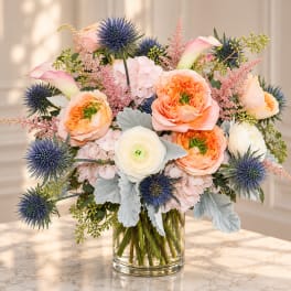 Peach and white flowers in a clear glass vase with blue thistles