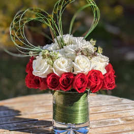 Round arrangement of red and white roses with white blooms and tall green loops in a clear glass vase.