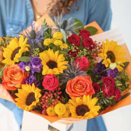 Mixed bouquet with sunflowers, roses, and purple blooms wrapped in orange paper