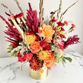 Vibrant arrangement of orange roses, red carnations and yellow protea in a gold cylinder vase