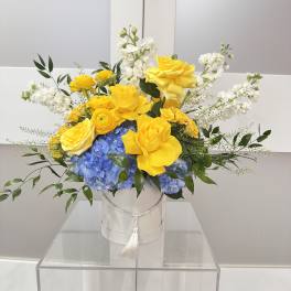 Yellow roses and blue hydrangeas in a white hatbox with white flowers.