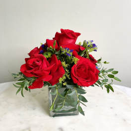 Red roses arranged in a square glass vase with greenery and small purple accents