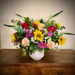 Colorful mixed arrangement with sunflowers, roses, and daisies in a white ceramic vase on a wooden table.