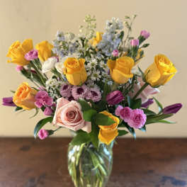 Mixed bouquet of yellow roses, pink roses, purple tulips, and small white daisies in a clear glass vase