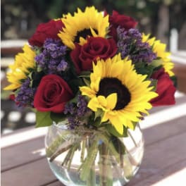 Round glass vase with yellow sunflowers, red roses, and small purple flowers in a compact bouquet
