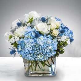 Low glass vase of blue hydrangeas, white roses, and small accent blooms in blue and white.